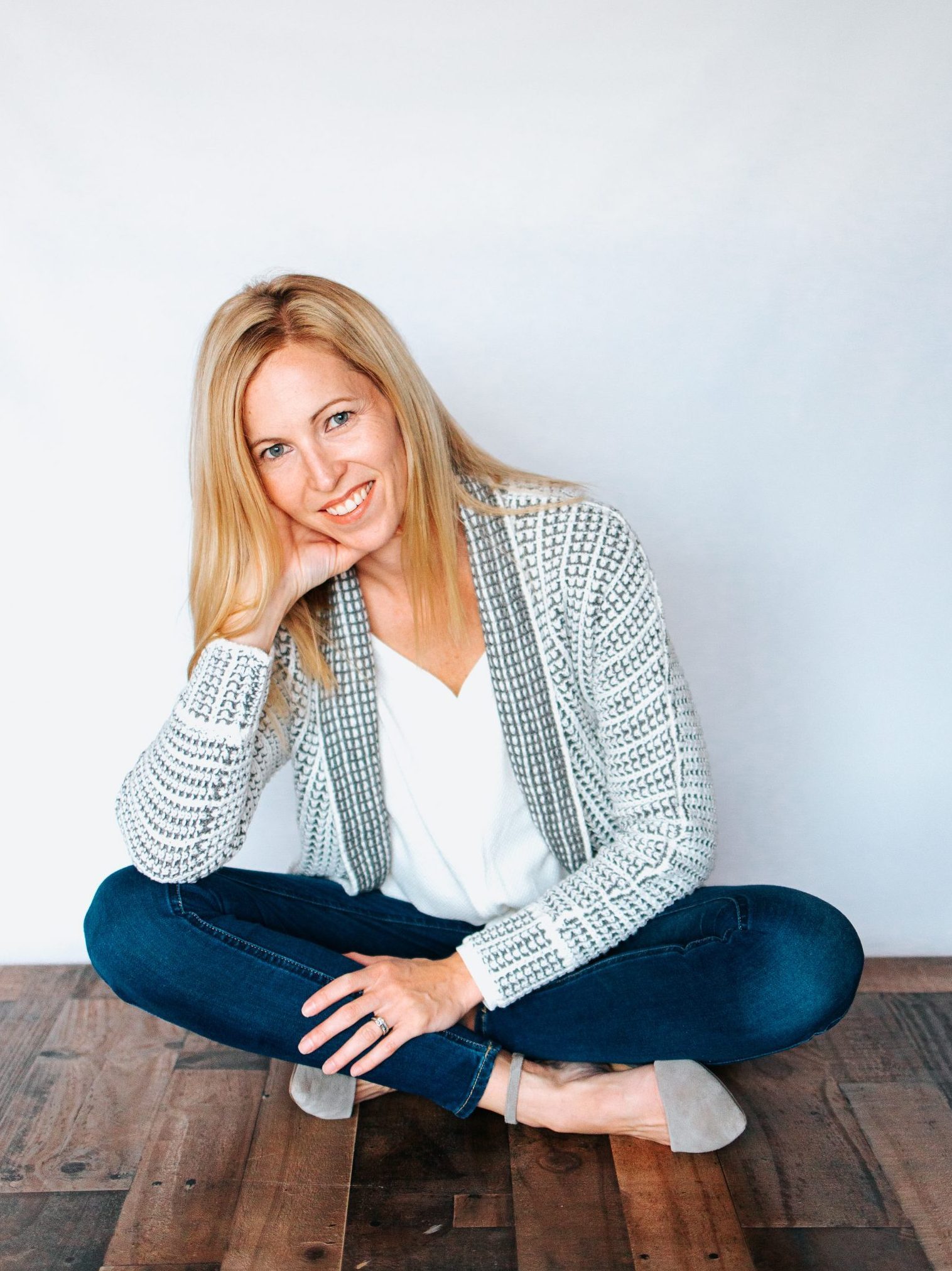 Kerri sitting on the floor in a blue cardigan and jeans, legs crossed, looking at the camera